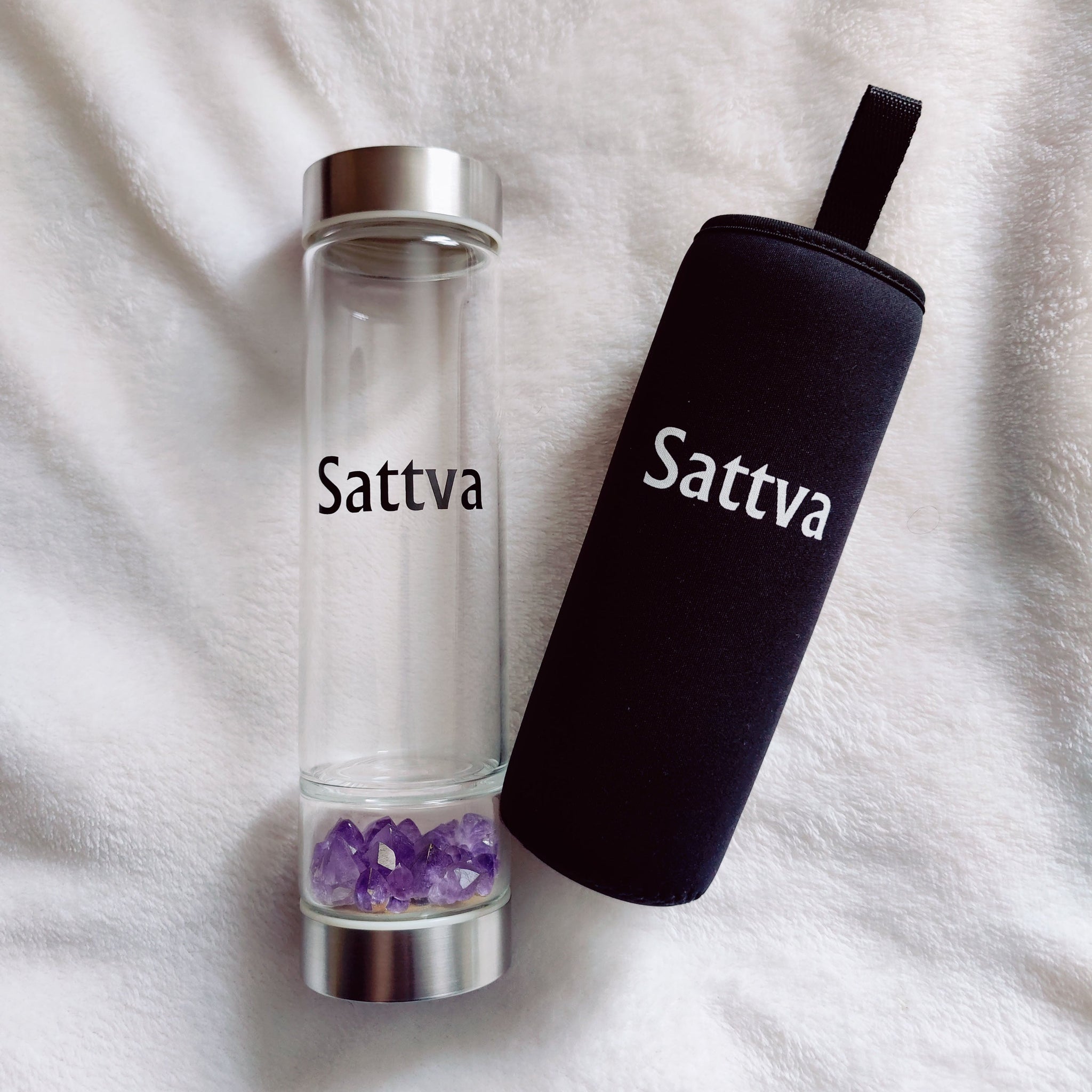 Clear glass water bottle with silver cap and black sleeve on a white fabric background, featuring 'Sattva' branding.