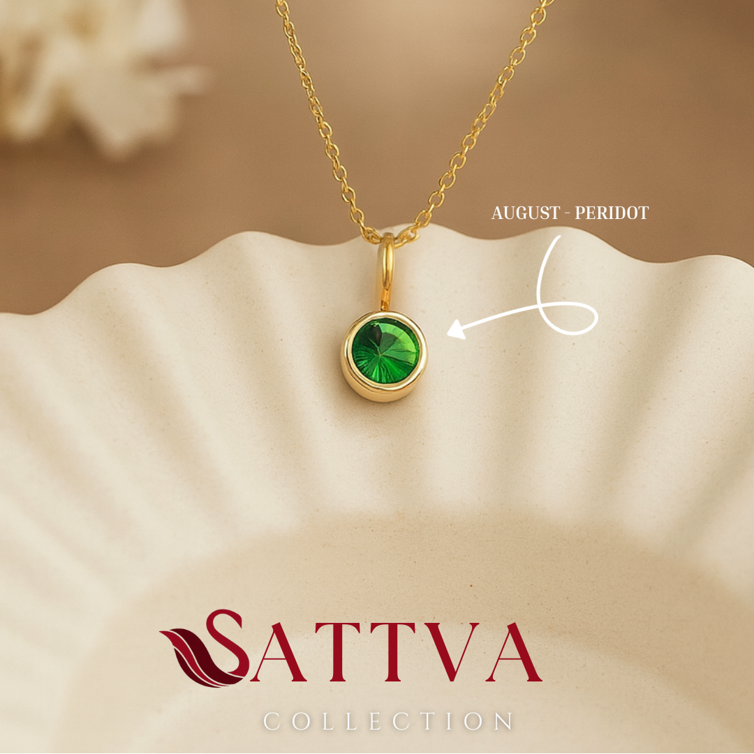 Gold necklace with a green gemstone pendant on a beige surface, featuring the Sattva Collection logo.