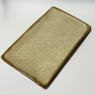 A rectangular ceramic serving platter with rounded corners, featuring a glossy brown border and a raised cream-colored mandala pattern on one half, set against a plain white background.