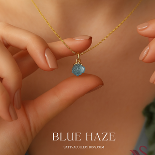 Gold necklace with a blue pendant held by a hand, brand 'Sativa Collections' visible