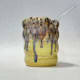 A ceramic cup with a yellow glazed base and dripping brown, purple, and blue glaze on the rim, featuring a clear glass straw inserted on the right side, photographed against a neutral light gray background.