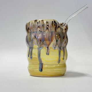 A ceramic cup with a yellow glazed base and dripping brown, purple, and blue glaze on the rim, featuring a clear glass straw inserted on the right side, photographed against a neutral light gray background.