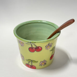 A light yellow ceramic cup decorated with red cherries and small pink flowers, with a green glazed interior and a dark brown wooden spoon resting inside, set against a plain light gray background.