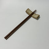 A pair of dark brown wooden chopsticks resting on a small beige ceramic chopstick holder against a plain light gray background.