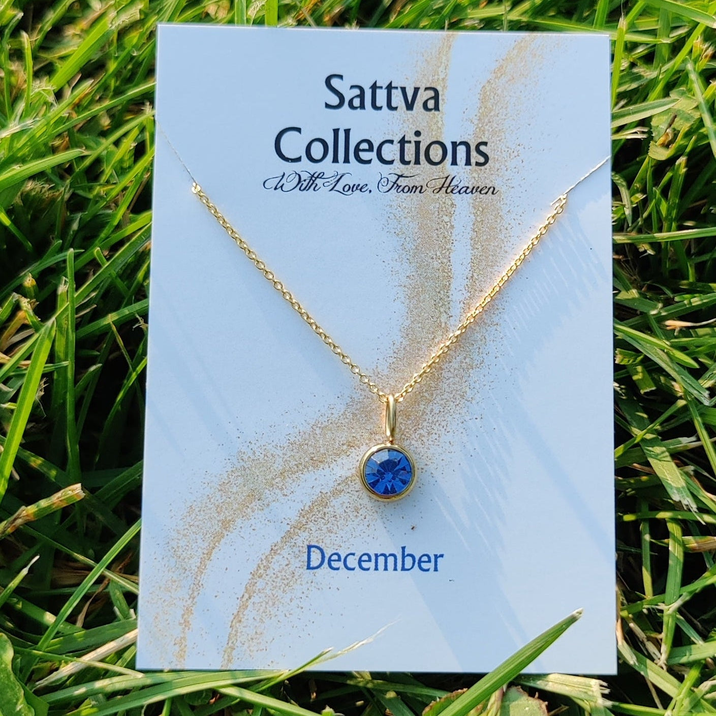 Gold necklace with a blue gemstone on a 'Sattva Collections' card in grass