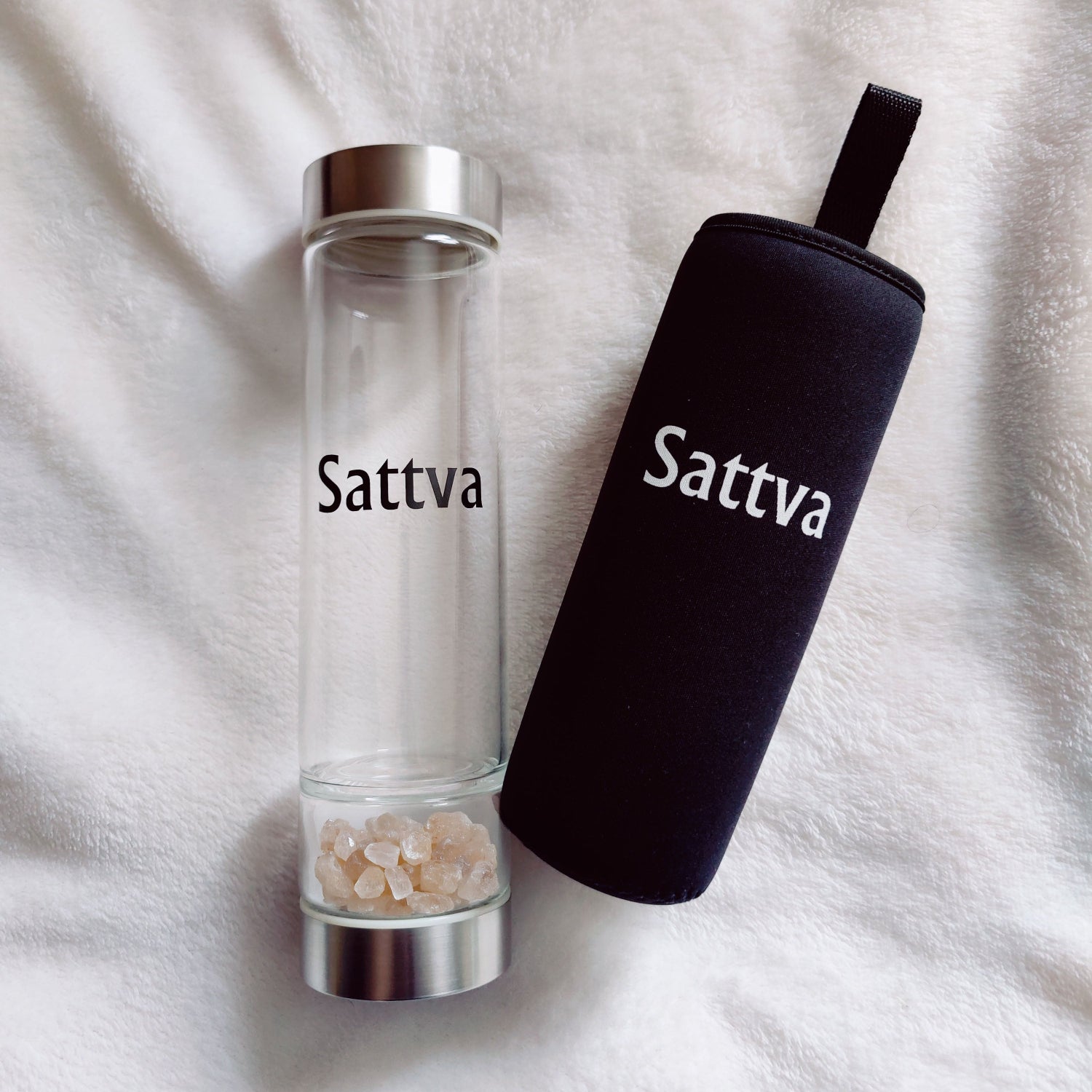 Clear glass bottle with silver cap and black sleeve labeled 'Sattva' on a white fabric background