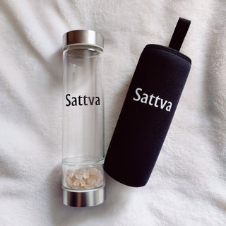 Clear glass bottle with silver cap and black sleeve labeled 'Sattva' on a white fabric background