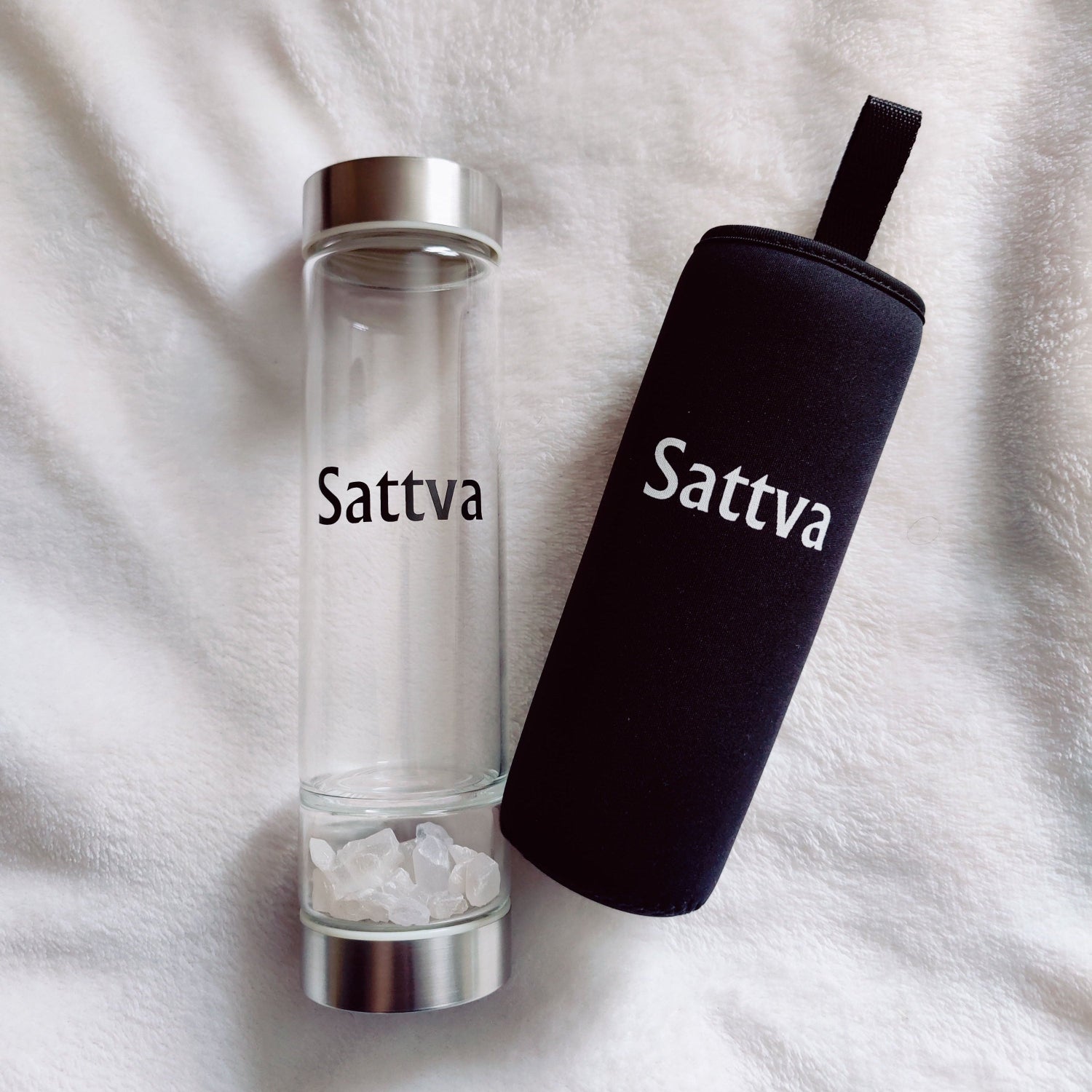Clear glass bottle with silver cap and black sleeve labeled 'Sattva' on a white fabric background