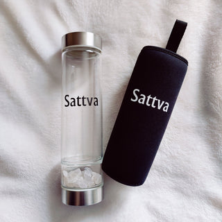 Clear glass bottle with silver cap and black sleeve labeled 'Sattva' on a white fabric background