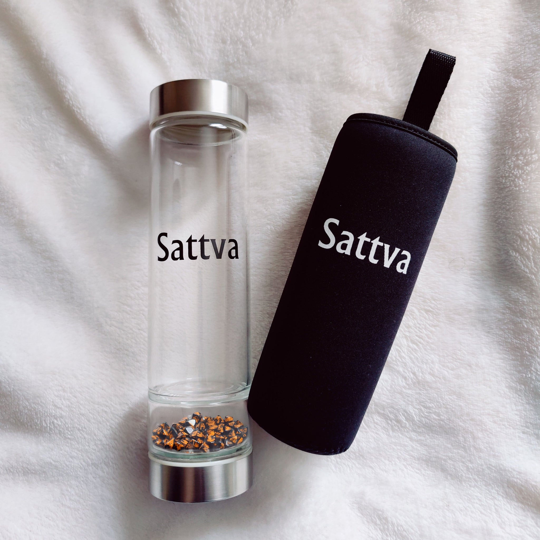 Clear glass bottle with silver cap and black sleeve labeled 'Sattva' on a white fabric background