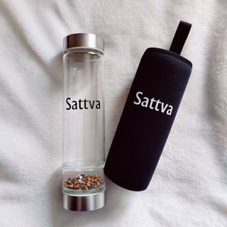 Clear glass bottle with silver cap and black sleeve labeled 'Sattva' on a white fabric background