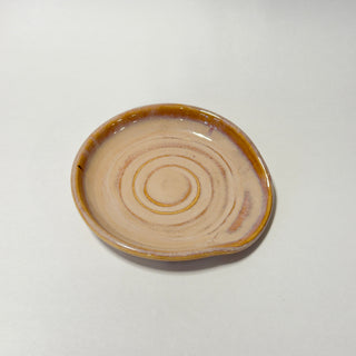 A small handmade ceramic dish with a glossy finish, featuring concentric circular patterns in a light beige glaze with a warm brown rim, set against a plain light gray background.