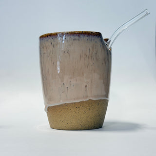 A handmade ceramic mug with a textured, speckled finish, featuring a glossy brown rim, a white drippy glaze detail, and a clear glass straw inserted into the side.