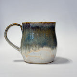 A handmade ceramic mug with a rounded body and a loop handle, featuring a glazed finish with a gradient of dark greeb, brown, and beige on a light gray background.