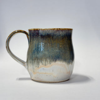 A handmade ceramic mug with a rounded body and a loop handle, featuring a glazed finish with a gradient of dark greeb, brown, and beige on a light gray background.