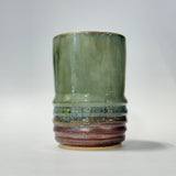 A handmade ceramic cup with a textured, ribbed base and a smooth, flared upper section, featuring a green and blue glaze that transitions into a rustic brown and terracotta pattern at the bottom, set against a plain light gray background.