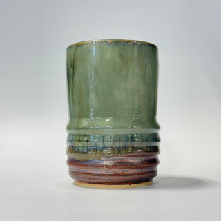 A handmade ceramic cup with a textured, ribbed base and a smooth, flared upper section, featuring a green and blue glaze that transitions into a rustic brown and terracotta pattern at the bottom, set against a plain light gray background.