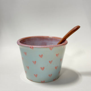 A light blue ceramic cup with a pink glazed rim and scattered pink hearts, containing a wooden spoon, set against a plain light gray background.