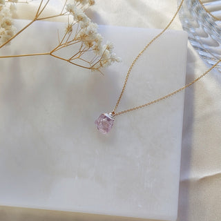 Gold necklace with a pink gemstone pendant on a white surface with dried flowers.