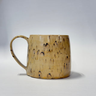 A handmade ceramic mug with a textured, organic splatter pattern in beige and brown, set against a neutral light gray background.