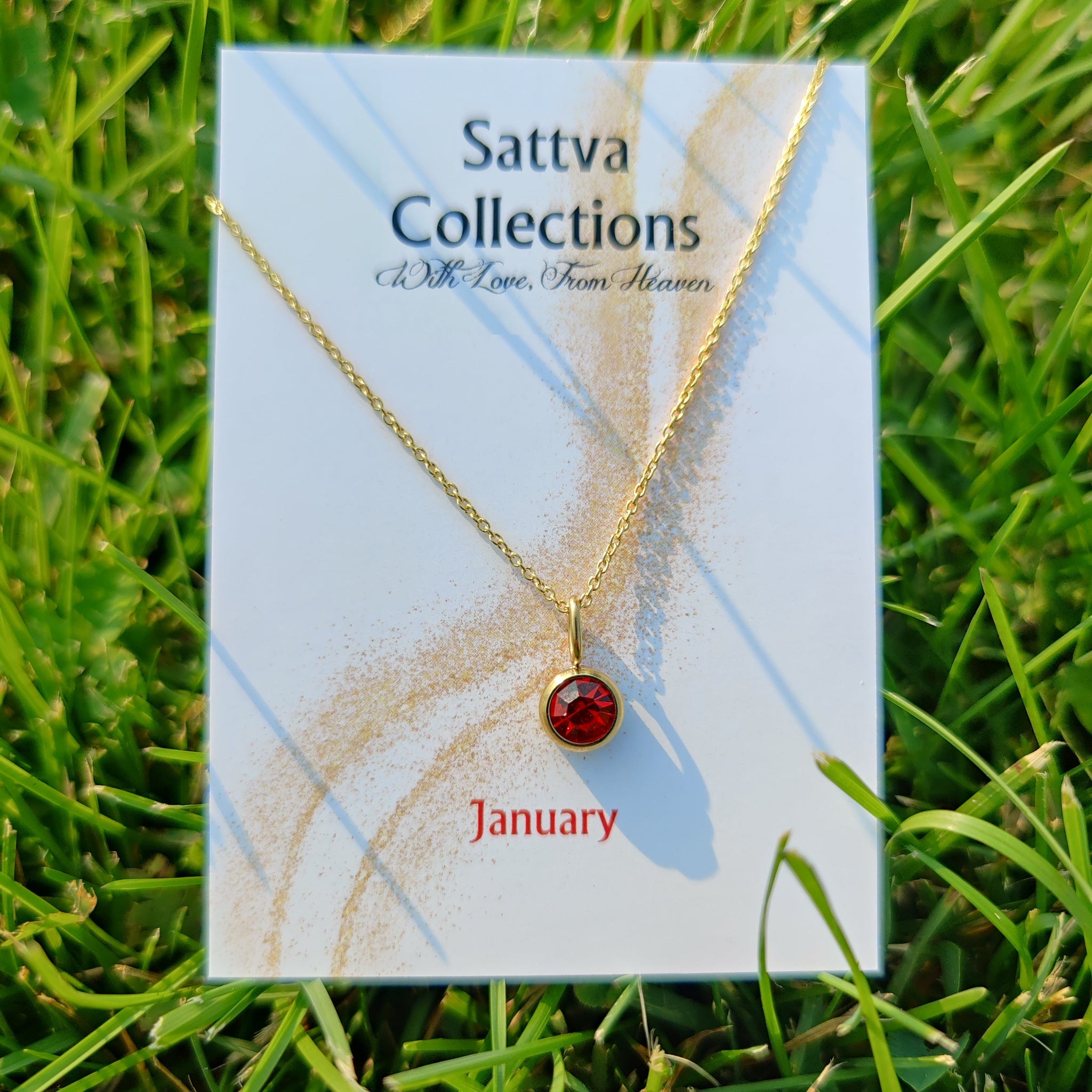 Gold necklace with a red gemstone on a 'Sattva Collections' card in grass