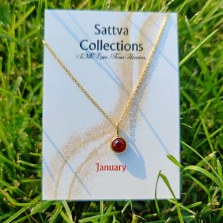 Gold necklace with a red gemstone on a 'Sattva Collections' card in grass