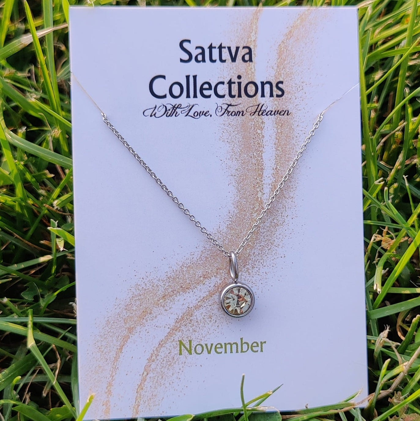 Necklace on a 'Sattva Collections' card with 'November' text, placed on grass.