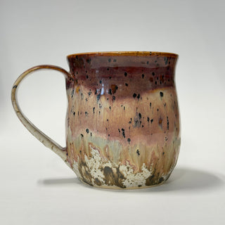 A handmade ceramic mug with a rustic, hand-painted glaze featuring a gradient of brown, beige, and pink with black speckles, set against a plain light gray background.