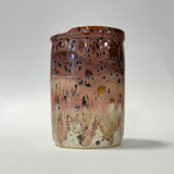 A handmade ceramic jar with a rustic, dripping glaze finish. The glaze transitions from a warm reddish-brown at the top to a dusty pink and cream at the bottom, heavily speckled with dark brown and black mineral flecks.