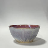 A handmade ceramic bowl with a textured, crackled glaze transitioning from a dusty lavender-purple on the outside to a warm reddish-brown on the inside and rim, set against a neutral gray studio background.