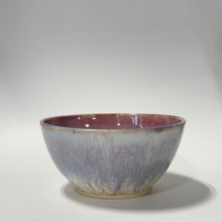 A handmade ceramic bowl with a textured, crackled glaze transitioning from a dusty lavender-purple on the outside to a warm reddish-brown on the inside and rim, set against a neutral gray studio background.