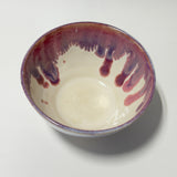 A handmade ceramic bowl with a glossy off-white interior and a rim decorated with dripping purple and reddish-brown glaze, shown from a top-down angle on a light gray background.