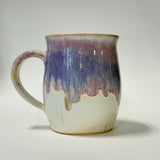 A handmade ceramic mug with a dripping glaze design, featuring a white base, a purple and blue middle section, and a brown rim, set against a plain light gray background.