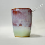 A handmade ceramic cup with a marbled glaze transitioning from a reddish-brown top to lavender-purple in the middle and a pale mint green base. A clear glass straw is inserted on the right side, set against a plain light gray background.