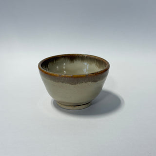 A handmade ceramic bowl with a glossy glaze, featuring a gradient transition from a warm beige base to a dark brown rim, set against a neutral gray studio background.