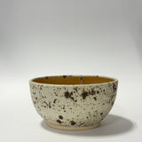A handmade ceramic bowl with a speckled cream exterior and a warm mustard-yellow glazed interior, photographed on a neutral light gray background.