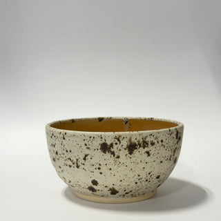 A handmade ceramic bowl with a speckled cream exterior and a warm mustard-yellow glazed interior, photographed on a neutral light gray background.