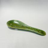 A handmade ceramic spoon rest with a glossy green glaze and speckled brown details, photographed on a neutral light gray background.