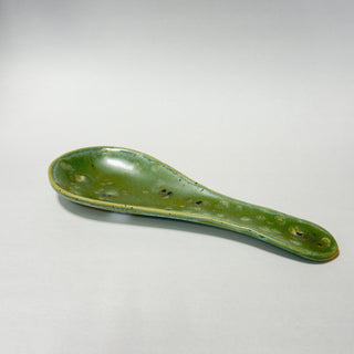 A handmade ceramic spoon rest with a glossy green glaze and speckled brown details, photographed on a neutral light gray background.