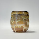 A handmade ceramic cup with a rounded shape, featuring a glossy glaze with a drip pattern in shades of brown, beige, and subtle hints of blue-green, set against a plain light gray background.
