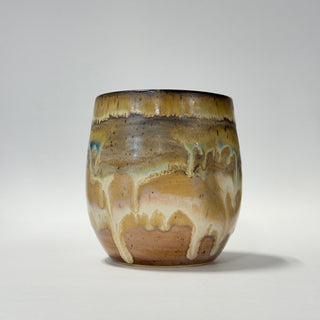 A handmade ceramic cup with a rounded shape, featuring a glossy glaze with a drip pattern in shades of brown, beige, and subtle hints of blue-green, set against a plain light gray background.
