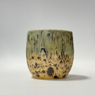 A handmade ceramic cup with a textured, mottled glaze transitioning from olive green at the top to a speckled tan and brown at the bottom, set against a neutral gray background.