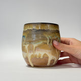 A handmade ceramic tumbler with a brown and beige drip glaze, held by a person's hand against a neutral gray background.