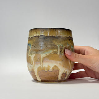 A handmade ceramic tumbler with a brown and beige drip glaze, held by a person's hand against a neutral gray background.