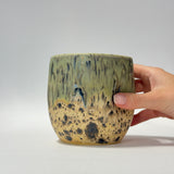 A ceramic cup with a textured, mottled glaze in olive green, beige, and black, held by a hand on the right side against a neutral gray background.