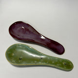 Two ceramic spoon rest with textured glazes, one in deep burgundy and the other in olive green, displayed diagonally on a light gray background.
