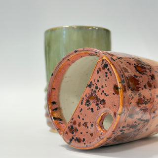 Two handmade ceramic vessels on a white studio background: a foreground planter with a terracotta-brown speckled glaze and a handle cutout, and a tall vase behind it with a muted green glaze.