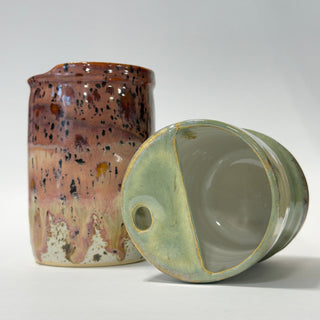 Two handmade ceramic vessels on a light gray studio background. One tall cylindrical jar has a brown, pink, and black speckled glaze, while the matching lidded container lies on its side showing a pale green-gray glazed interior and rim.