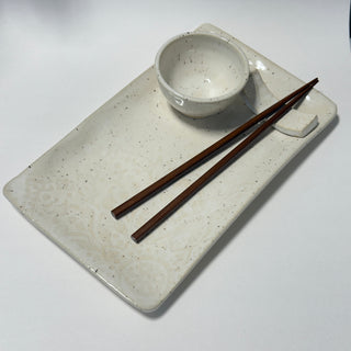 An off-white speckled ceramic rectangular serving tray with a small matching bowl and a pair of dark brown chopsticks, photographed on a plain white background.