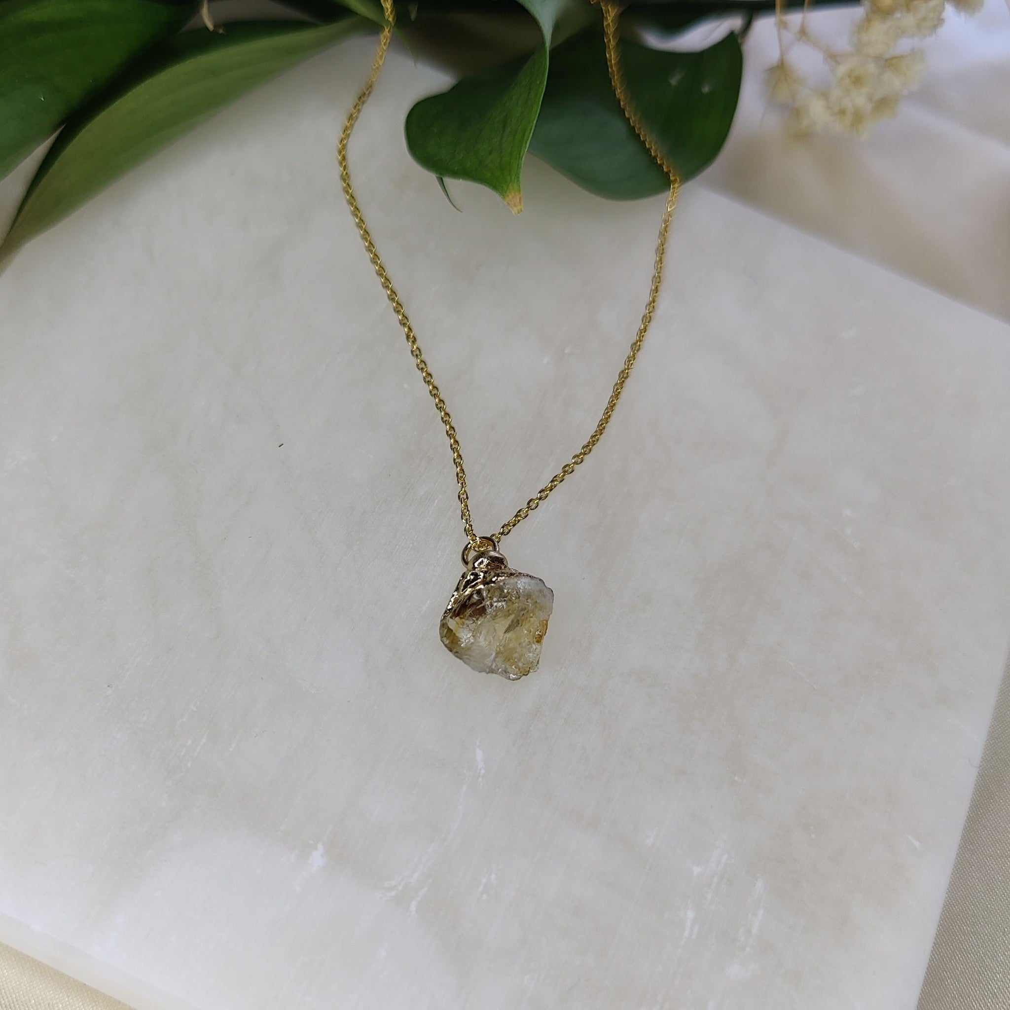 Gold necklace with a pendant on a white marble surface with green leaves and flowers.